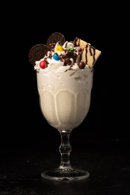 Siyah arka planda şeffaf bir bardakta tatlı, waffle, kurabiye ve kremayla süslenmiş klasik milkshake.