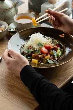 Kız yemek çubuklarıyla tavuk, havuç, ızgara mısır, peynir ve vişneli domatesli baharatlı Asya sebze salatası yiyor. Bir Asya restoranında öğle yemeği. Sığ alan derinliği, bulanık arkaplan.