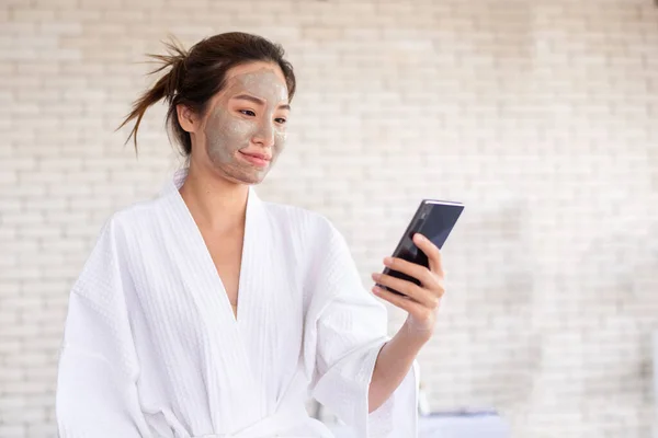 Yüzünde kil maske olan akıllı telefon kullanan Asyalı kadın.