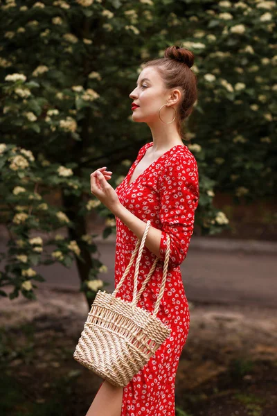 a model in a city park holds a straw bag. stylish summer wear. flowering trees in the background