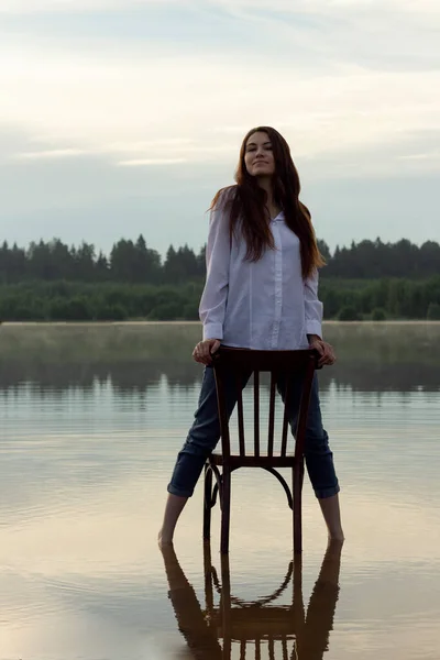 The girl is standing on the water in a white shirt and jeans, holding the back of a chair. A girl with an old chair in the middle of the lake.