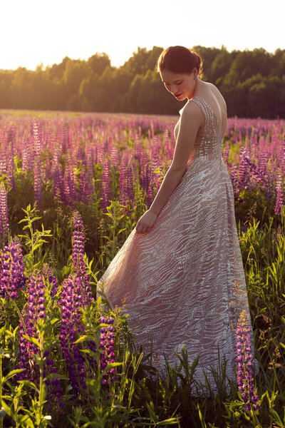 A girl stands with her back in an evening dress on a field of lupins. Open back on an evening dress.