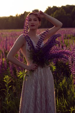 Bir buket lupinle güzel bir kız, gece elbisesi içinde, bir çiçek çayırında duruyor..