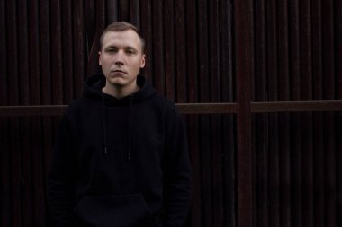 Paslı borulardan bir duvara karşı siyah kapüşonlu sarışın bir adam. Siyah ceketli, açık renk yakışıklı bir adamın portresi..