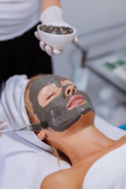 Kozmetik uzmanı güzellik kliniğinde, kaplıca salonunda genç ve güzel bir kadının yüzüne kilden maske takıyor. Beyaz Avrupalı kadın için yüz bakımı prosedürü uygulayan güzellik uzmanı