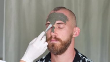 Kozmetik uzmanı, güzellik kliniğinde, güzellik salonunda genç yakışıklıların yüzüne kilden maske takıyor. Beyaz tenli Avrupalı erkeklere yüz bakımı yapan güzellik uzmanı.  