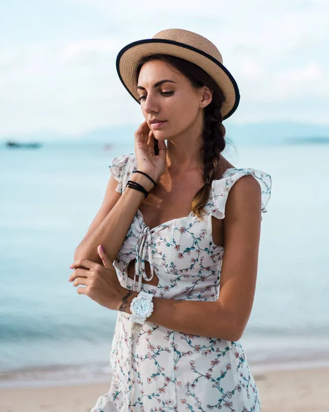 Positive calm woman in light summer dress,  straw hat and watches on tropical beach, summer vibes, vacation needed. Sunset light, warm weather. Close fashion portrait.