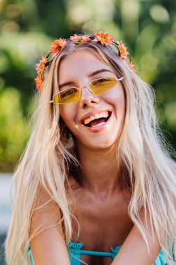 Sarı renkli güneş gözlüklü, mavi seksi bikinili, kafasında çelenk olan, yaz havasında, şık giyinen, uzun saçlı genç bir kadının portresi. Yüzme havuzunda tatilde olan bir kadın..