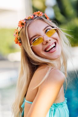 Sarı renkli güneş gözlüklü, mavi seksi bikinili, kafasında çelenk olan, yaz havasında, şık giyinen, uzun saçlı genç bir kadının portresi. Yüzme havuzunda tatilde olan bir kadın..