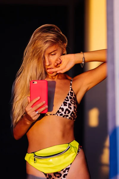 暖かい日没の光でヒョウビキニの若いフィットスリムブロガー女性の明るいカラフルなスタイリッシュな肖像画は鏡で写真自撮りを取ります ネオンカラー 酸黄緑色のウエストバッグとピンクの電話ケース — ストック写真