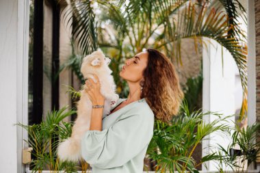 Avrupalı kıvırcık tenli kadının açık hava portresi mutlu köpekli pomeranian spitz tutuyor. Elinde gümüş bilezik var. Güzel beyaz tüylü köpek yavrusuyla vakit geçirmek. Evcil hayvanlarla mutlu anlar. Egzotik arkaplan.