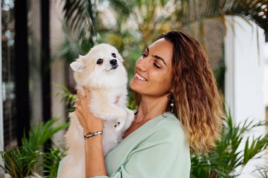 Avrupalı kıvırcık tenli kadının açık hava portresi mutlu köpekli pomeranian spitz tutuyor. Elinde gümüş bilezik var. Güzel beyaz tüylü köpek yavrusuyla vakit geçirmek. Evcil hayvanlarla mutlu anlar. Egzotik arkaplan.