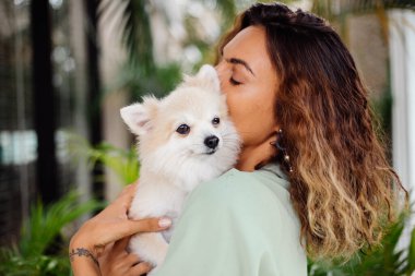 Avrupalı kıvırcık tenli kadının açık hava portresi mutlu köpekli pomeranian spitz tutuyor. Elinde gümüş bilezik var. Güzel beyaz tüylü köpek yavrusuyla vakit geçirmek. Evcil hayvanlarla mutlu anlar. Egzotik arkaplan.