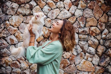 Romantik yaz elbisesi içindeki genç Avrupalı kadın, bilezik, tuğla duvarın arkasında köşkün dışında sevimli pomeranya köpek salyası tutuyor..