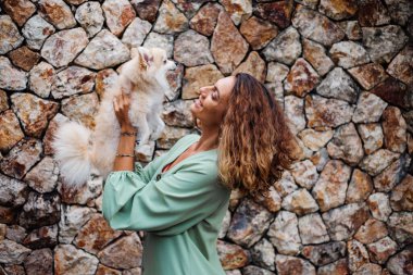 Romantik yaz elbisesi içindeki genç Avrupalı kadın, bilezik, tuğla duvarın arkasında köşkün dışında sevimli pomeranya köpek salyası tutuyor..