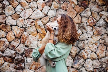 Romantik yaz elbisesi içindeki genç Avrupalı kadın, bilezik, tuğla duvarın arkasında köşkün dışında sevimli pomeranya köpek salyası tutuyor..