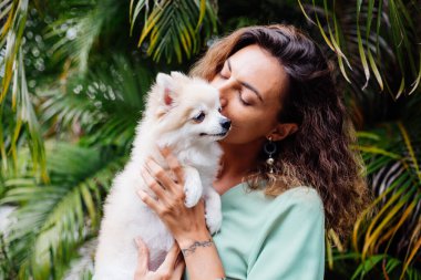 Avrupalı kıvırcık tenli kadının açık hava portresi mutlu köpekli pomeranian spitz tutuyor. Elinde gümüş bilezik var. Güzel beyaz tüylü köpek yavrusuyla vakit geçirmek. Evcil hayvanlarla mutlu anlar. Egzotik arkaplan.