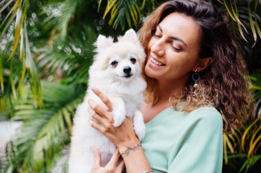 Avrupalı kıvırcık tenli kadının açık hava portresi mutlu köpekli pomeranian spitz tutuyor. Elinde gümüş bilezik var. Güzel beyaz tüylü köpek yavrusuyla vakit geçirmek. Evcil hayvanlarla mutlu anlar. Egzotik arkaplan.