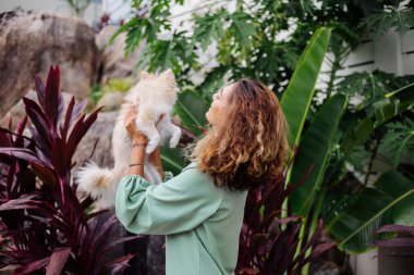 Avrupalı kıvırcık tenli kadının açık hava portresi mutlu köpekli pomeranian spitz tutuyor. Elinde gümüş bilezik var. Güzel beyaz tüylü köpek yavrusuyla vakit geçirmek. Evcil hayvanlarla mutlu anlar. Egzotik arkaplan.