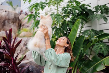 Avrupalı kıvırcık tenli kadının açık hava portresi mutlu köpekli pomeranian spitz tutuyor. Elinde gümüş bilezik var. Güzel beyaz tüylü köpek yavrusuyla vakit geçirmek. Evcil hayvanlarla mutlu anlar. Egzotik arkaplan.