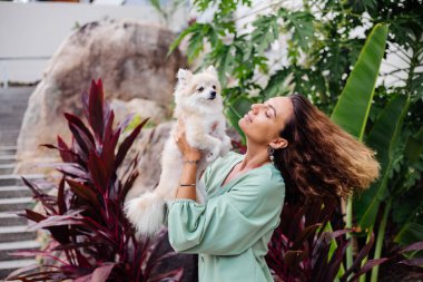 Avrupalı kıvırcık tenli kadının açık hava portresi mutlu köpekli pomeranian spitz tutuyor. Elinde gümüş bilezik var. Güzel beyaz tüylü köpek yavrusuyla vakit geçirmek. Evcil hayvanlarla mutlu anlar. Egzotik arkaplan.