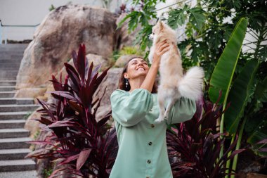 Avrupalı kıvırcık tenli kadının açık hava portresi mutlu köpekli pomeranian spitz tutuyor. Elinde gümüş bilezik var. Güzel beyaz tüylü köpek yavrusuyla vakit geçirmek. Evcil hayvanlarla mutlu anlar. Egzotik arkaplan.