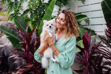Avrupalı kıvırcık tenli kadının açık hava portresi mutlu köpekli pomeranian spitz tutuyor. Elinde gümüş bilezik var. Güzel beyaz tüylü köpek yavrusuyla vakit geçirmek. Evcil hayvanlarla mutlu anlar. Egzotik arkaplan.
