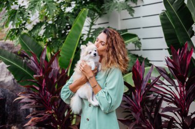 Avrupalı kıvırcık tenli kadının açık hava portresi mutlu köpekli pomeranian spitz tutuyor. Elinde gümüş bilezik var. Güzel beyaz tüylü köpek yavrusuyla vakit geçirmek. Evcil hayvanlarla mutlu anlar. Egzotik arkaplan.