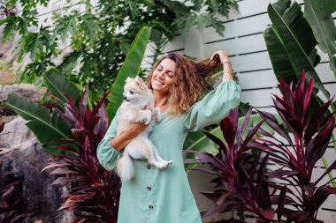 Avrupalı kıvırcık tenli kadının açık hava portresi mutlu köpekli pomeranian spitz tutuyor. Elinde gümüş bilezik var. Güzel beyaz tüylü köpek yavrusuyla vakit geçirmek. Evcil hayvanlarla mutlu anlar. Egzotik arkaplan.