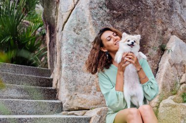 Avrupalı kıvırcık tenli kadının açık hava portresi mutlu köpekli pomeranian spitz tutuyor. Elinde gümüş bilezik var. Güzel beyaz tüylü köpek yavrusuyla vakit geçirmek. Evcil hayvanlarla mutlu anlar. Egzotik arkaplan.