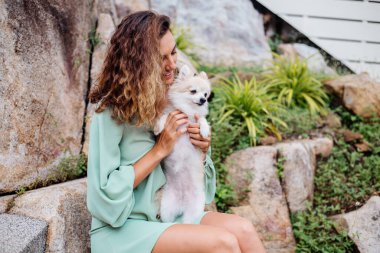 Avrupalı kıvırcık tenli kadının açık hava portresi mutlu köpekli pomeranian spitz tutuyor. Elinde gümüş bilezik var. Güzel beyaz tüylü köpek yavrusuyla vakit geçirmek. Evcil hayvanlarla mutlu anlar. Egzotik arkaplan.