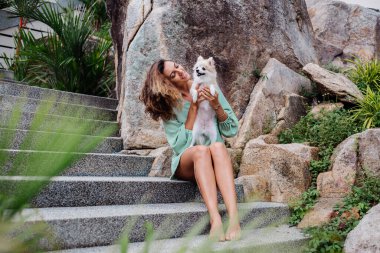 Avrupalı kıvırcık tenli kadının açık hava portresi mutlu köpekli pomeranian spitz tutuyor. Elinde gümüş bilezik var. Güzel beyaz tüylü köpek yavrusuyla vakit geçirmek. Evcil hayvanlarla mutlu anlar. Egzotik arkaplan.