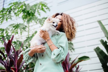 Avrupalı kıvırcık tenli kadının açık hava portresi mutlu köpekli pomeranian spitz tutuyor. Elinde gümüş bilezik var. Güzel beyaz tüylü köpek yavrusuyla vakit geçirmek. Evcil hayvanlarla mutlu anlar. Egzotik arkaplan.