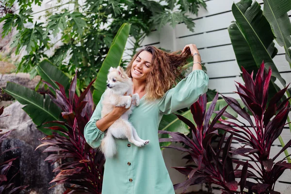 Avrupalı kıvırcık tenli kadının açık hava portresi mutlu köpekli pomeranian spitz tutuyor. Elinde gümüş bilezik var. Güzel beyaz tüylü köpek yavrusuyla vakit geçirmek. Evcil hayvanlarla mutlu anlar. Egzotik arkaplan.