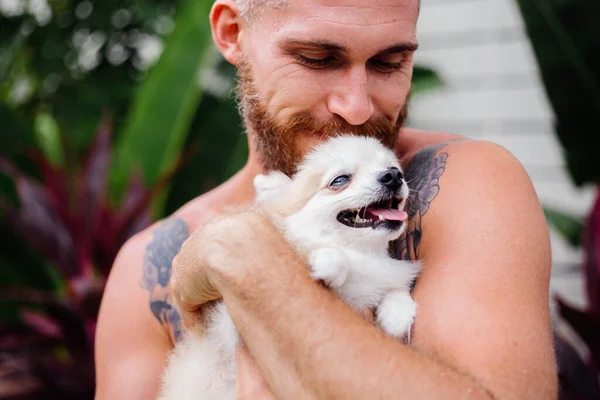 Young handsome bearded brutal tattooed happy man holds pomeranian spitz, playing with lovely pet. Male spending time outside in park with cute fluffy dog puppy. Tropical exotic background.