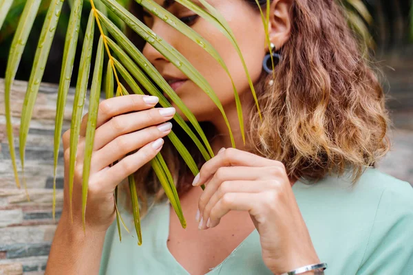 Close outdoor portrait of tanned european woman with short hair in round jewerly earings with palm leaf tropical background.