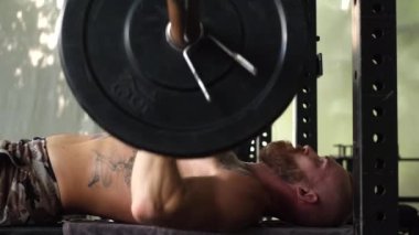 Spor salonunda sakallı, güçlü bir adam dövmesi var. Halter egzersizi yapan kaslı vücutçu. Genç sporcu halter yarışmalarına katılıyor..      