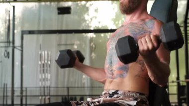 Genç, kaslı, güçlü, kaslı, sakallı Avrupalı adam omuzlarında dambıllarla sıkı egzersiz yapıyor.    