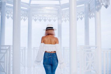 Havalı kolları, kotu ve klasik şapkası olan, sütunlu güzel beyaz kemerli, arka planda mavi deniz olan modaya uygun bir kadının arka planı..