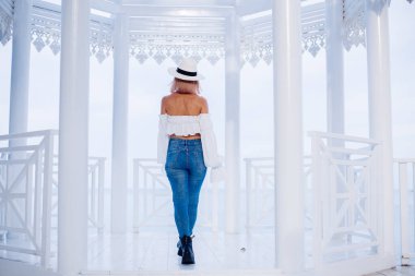 Havalı kolları, kotu ve klasik şapkası olan, sütunlu güzel beyaz kemerli, arka planda mavi deniz olan modaya uygun bir kadının arka planı..