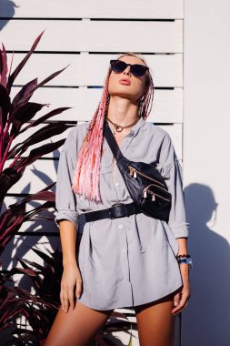 Gri uzun gömlekli, pembe mor örgülü, aksesuarlı, güneş gözlüklü, siyah bel çantalı moda saatli, elinde tropikal arka plan ve beyaz ahşap duvarlı genç Avrupalı stil kadın..