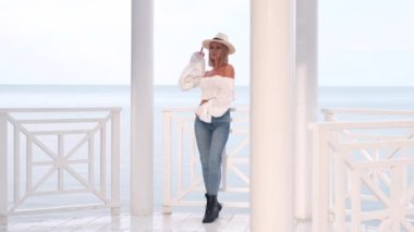 Havalı kolları, kotu ve güzel beyaz kemerli klasik şapkası olan, sütunlu, arka planda mavi denizi olan şık bir kadın..    