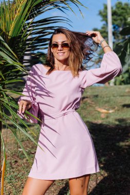 Romantik pembe elbiseli, yuvarlak küpeli, gümüş bilezik ve güneş gözlüklü, kır çiçekli, tropikal egzotik arka planda, beyaz tenli genç bir kadının portresi. Sakin güzel kadın parkta güneşleniyor.