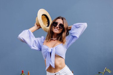 Mor renkli, şık bir kadın uzun kollu, beyaz şort, siyah güneş gözlüğü, hasır şapka ve inci kolye, izole, mavi gri duvar, doğal gün ışığı..