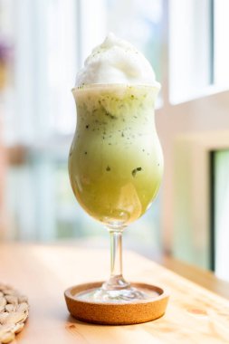 Buzlu kibrit Japon yeşil çayı, beyaz süt köpüğü ve güzel bir bardakla birlikte, yaz kafeteryasındaki ahşap bir masaya yerleştirilmiş, doğal gün ışığı..