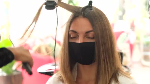一个女理发师用熨斗把客户的角蛋白头发拉直 这个女孩戴着黑色的口罩坐在那里 社会距离 保护措施 检疫结束了 — 图库视频影像