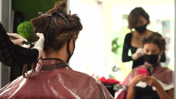 一个女理发师使客户的角蛋白头发直 这个女孩穿着黑色的医疗面罩坐在沙龙里 用吹风机吹风社会距离 保护措施 有电话的客户 — 图库视频影像