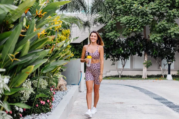 Fashion stylish young caucasian fit sporty woman in leopard cami top and  biker shorts outside holds protein shaker,  bottle of water, in hand. Female with abs after jogging thirsty for water.