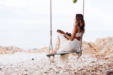 Romantik, genç, mutlu, sakin, Kafkas dövmeli, günbatımında salıncakta sallanan tropikal kayalık plajda gitar çalan yalnız bir kadın.  