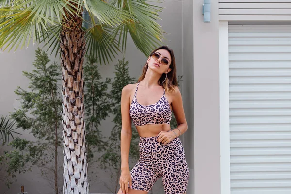 Fashion stylish fit tanned european woman in sunglasses, leopard cami top and biker shorts, outside villa near palm tree.